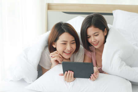 Asian young sisters lovely couple sitting on white bed and smiling together for Social online with laptop and smartphone in bedroom. Homosexual and asian Lesbian in relationship. Technology Conceptの写真素材