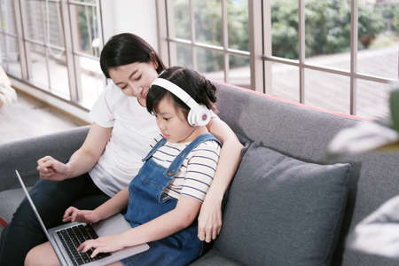 Smiling Asian mother and little girl child is relaxing with headphone and laptop for listening to online music or cartoon movie in social media on sofa in living room. Education and technology conceptの写真素材
