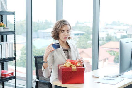Businesswoman holding credit card and surprise with red gift box at office computer desk. Valentine's Day, Christmas, birthday, shopping conceptの写真素材