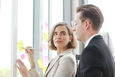 Successful businessman and businesswoman writing good ideas on sticky notes pasted on glass wall in modern office meeting.の写真素材