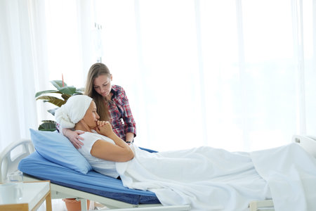 Caucasian daughter is Holding hands Elderly Mother in white headscarf is laying on bed in hospital after chemotherapy because she is suffering from cancer or Leukemia patient. focus on hands.の写真素材