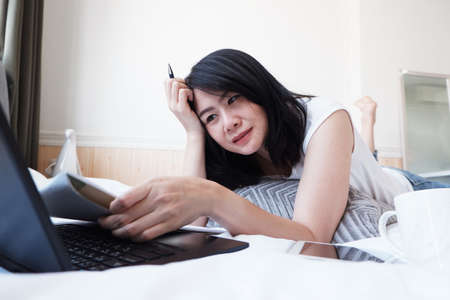 Smiling Asian businesswoman is writing and researching with laptop in bedroom on holiday. She is online working from home with technology during the Covid-19 outbreak concept.の写真素材