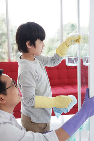 Smiling Asian father and little boy child is enjoying for cleaning spray glass cleaner and Wiping the glass together in Laundry room. Family time on holiday conceptの写真素材