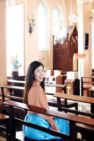 Asian woman is sitting on wooden chair and smiling for Pray to god in a Christian church for make a wish for hope and encouragement in life. Rituals and beliefs in Christian of Thailand concept.の写真素材