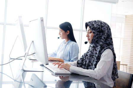 Smiling asian Muslim women is call center or secretary operator is wearing a headset and a microphone for consultant to customers. Technician Support staff for help resolve technical issues.の写真素材