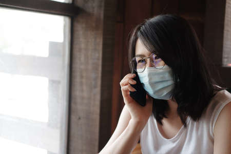 Asian woman is wearing mask for prevent covid-19 and calling on smartphone. Businesswoman is online working with laptop in living room. Work from home for Corona visus outbreak concept.の写真素材