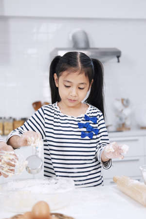 Smiling Asian little asian girl child is baking bakery cake and cookies on wooden table in kitchen....