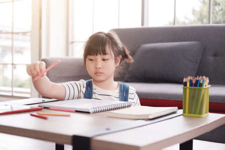 Smiling cute Asian little girl child is drawing and Painting with wooden colored pencils on paper in living room. She is online learning for Homeschool and educational conceptの写真素材