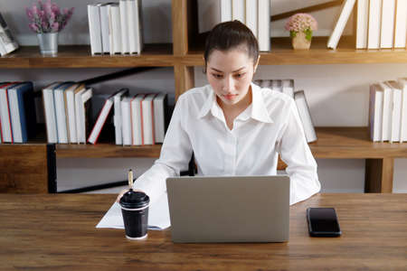 Asian businesswoman is working or online shopping with laptop and drinking hot coffee in workspace...
