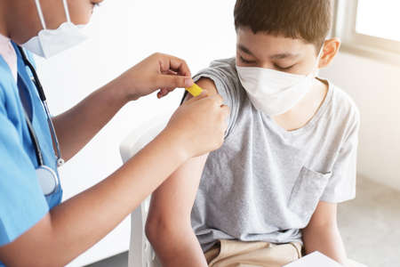 Profssional African doctor or nurse woman is vaccinating and cover plaster to Caucasian boy is wearing mask in the hospital. Vaccines for children to prevent COVID-19の写真素材