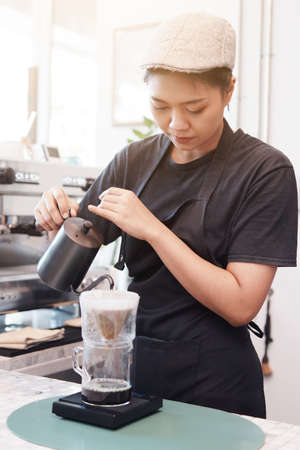 Smiling Asian barista young woman is wearing apron and pouring and craft a hot black coffee into cup for according to the customer's order at counter bar in coffee shop.の写真素材