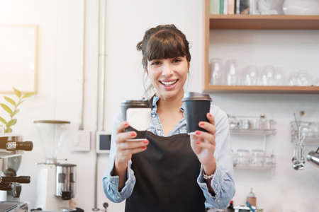 Smiling Caucasian Young barista woman is wearing apron and holding two cups of hot coffee at counter bar in cafe. Start up for Coffee shop and cafe business owner concept.の写真素材