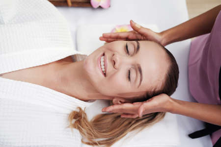 Asian Massage therapist woman is making traditional head and facial treatment massage to Caucasian female is relaxing and smilng on bed at alternative medicine healing spa Center in Thailand.の写真素材