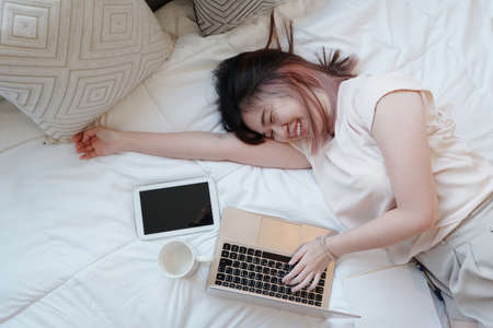 Smiling Asian businesswoman is relaxing and laying with laptop in bedroom on holiday. She is online working from home with technology during the Covod-19 outbreak concept.の写真素材