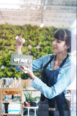 Smiling Caucasian Young barista woman or florist is wearing apron for setting open sign label on...