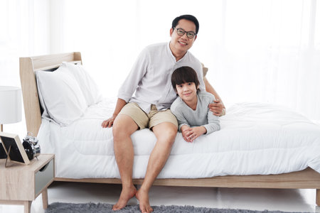 Smiling Asian cute little boy is relaxing and hugging with father in bedroom at home. Family love and child educational for homeschool on holiday concept.の写真素材