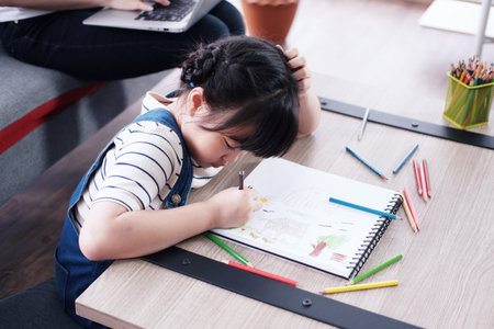Smiling cute Asian little girl child is drawing and Painting with wooden colored pencils on paper in living room. She is online learning for Homeschool and educational conceptの写真素材