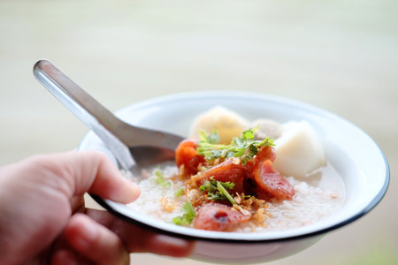 Woman hand holding rice porridge with pork and Fried Chinese sausage in white bowl.  Favorite food and tradition breakfast in Thailandの写真素材