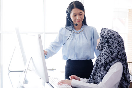Smiling Asian and Muslim women are colleagues and call center or secretary operator with headset and a microphone together. They are Consulting to solve problems about customers at officeの写真素材