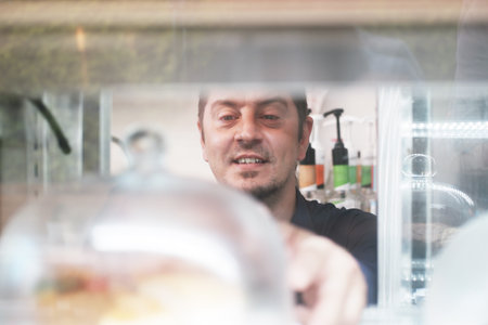 Smiling Caucasian barista and baker man is wearing apron open cake and bakery cabinet in coffee shop...