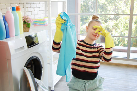 Smiling Caucasian young female is housekeeper wearing  yellow gloves and washing clothes. She cover...