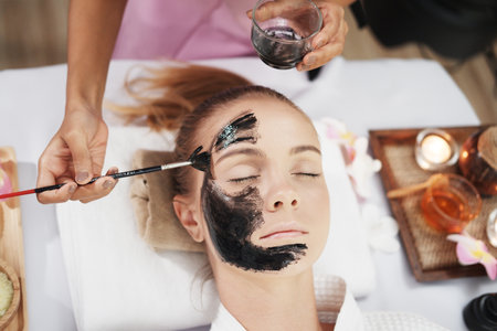 Close up on hand professional Asian Thai Massage therapist woman treatment black clay facial mask for caucasian female skin care on face at alternative medicine healing spa Center in Thailandの写真素材