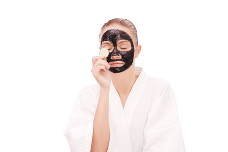 Smiling Caucasian teenager girl wearing bathrobe and holding sliced cucumber and relaxing facial mask black clay for skin face care in spa treatment isolated on white background with clipping pathの写真素材
