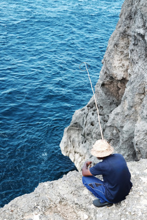 An elderly Balinese man wearing straw hat and fishing on the edge of a sea cliffの写真素材
