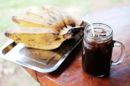 A drinking glass of iced black coffee and tropical banana on wooden tableの写真素材