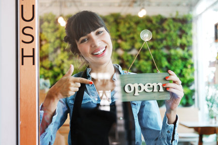 Smiling Caucasian barista woman or florist wearing black apron holding open sign label on glass door...