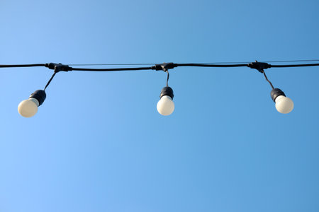 Vintage glowing light bulbs hang from a thin wire in the air and blue sky in bright day