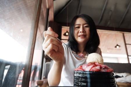 Smiling Asian woman eating shaved ice strawberry bingsu on wooden table in cafeの写真素材