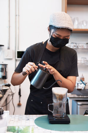 Professional Asian woman barista wearing mask and black apron pouring hot water into the coffee dripper in cafeの写真素材