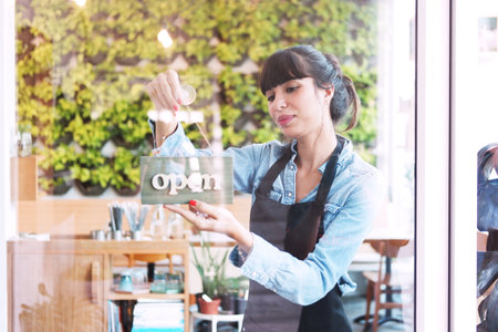 Smiling Caucasian barista businesswoman or florist wearing apron for setting open sign label on...