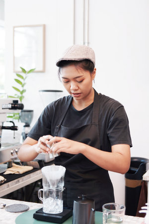 Professional Asian woman barista wearing black apron and pouring cocoa powder into the coffee dripper on counter bar in cafe or coffee shopの写真素材