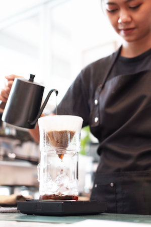 Professional Asian woman barista wearing black apron and pouring hot water into the coffee dripper...