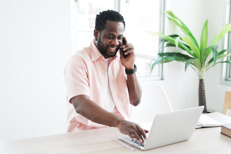 African American businessman using and talking smartphone for online working with laptop on desk in...