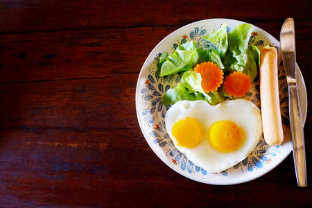 Fresh heart shaped fried eggs with organic vegetable salad and hot dog in white dish breakfast on wooden tableの写真素材