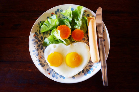 Top view of fresh heart shaped fried eggs with vegetable salad and hot dog in white dish breakfast on wooden tableの写真素材