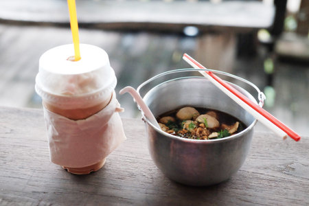 Thai boat noodles with chicken soup and pork balls in aluminum bowl with iced coffee in plastic...