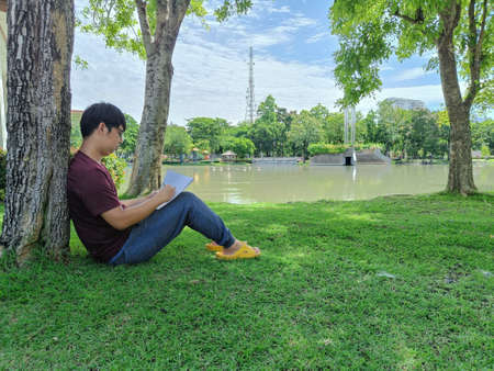 Young Asian man with black hair sits under a tree holding a book. Stress and anxiety. What are you...