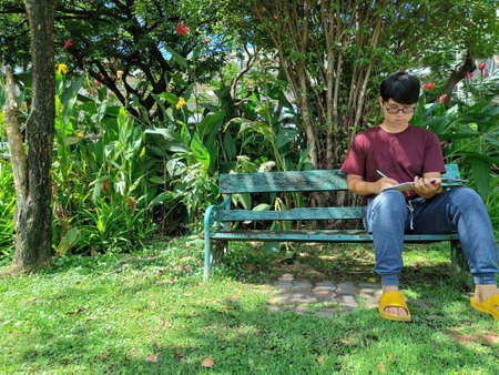 Young Asian man wearing glasses with black hair holding a book on a bench. happy and relax. What are thinking in the summer park cityの写真素材