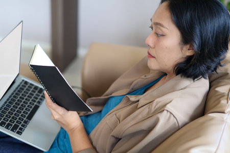 Close up view of a hand holding a notebook to view information while working on a laptop. Working from home, workspace.の写真素材