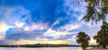 Sunset reflected in the river. Orange and pink clouds scatter. Calm evening river reflects the sky. Yellow and orange tones turn into purple shadows. Wide panoramic view of water and vegetation. Fresh water. Water quality.の写真素材