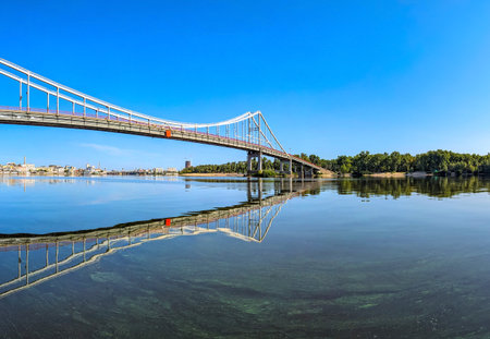 Pedestrian crossing suspension structures. Bridge over the Dnieper River with a wonderful panorama of blue sky and soft waves in the foreground. Wonderful zigzag reflection spans and support in the water.の写真素材