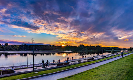 Orange sunset on the embankment. Perspective of the pedestrian zone near the river. People are calmly walking in the evening. Calm and peace. Beautiful colorful spectacle of clouds is reflected in the water of the river.の写真素材