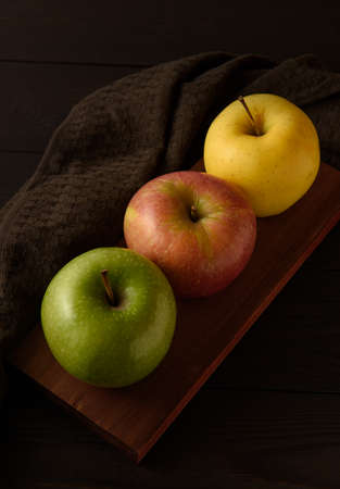 Three multicolored apples on a brown wooden board on a dark brown wooden background.の写真素材