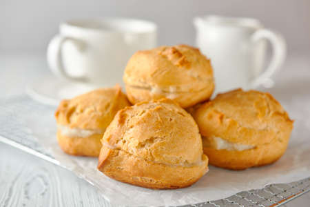 Homemade profiteroles on a metal grid and white paper on a white wooden background. Behind are a teacup and a milk jug.の写真素材