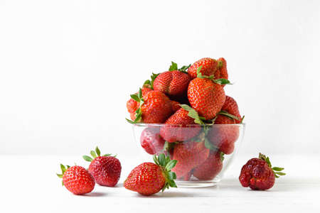 Fresh strawberries in a glass bowl on a white background. A few berries are lying on the wooden table. Close up, copy space.の写真素材