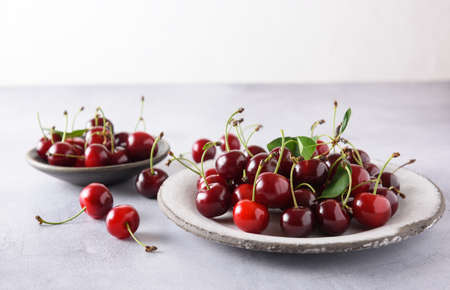 Fresh cherries on a white ceramic plate on a light gray background. In the background is a gray saucer with a cherry. Close up, copy space.の写真素材